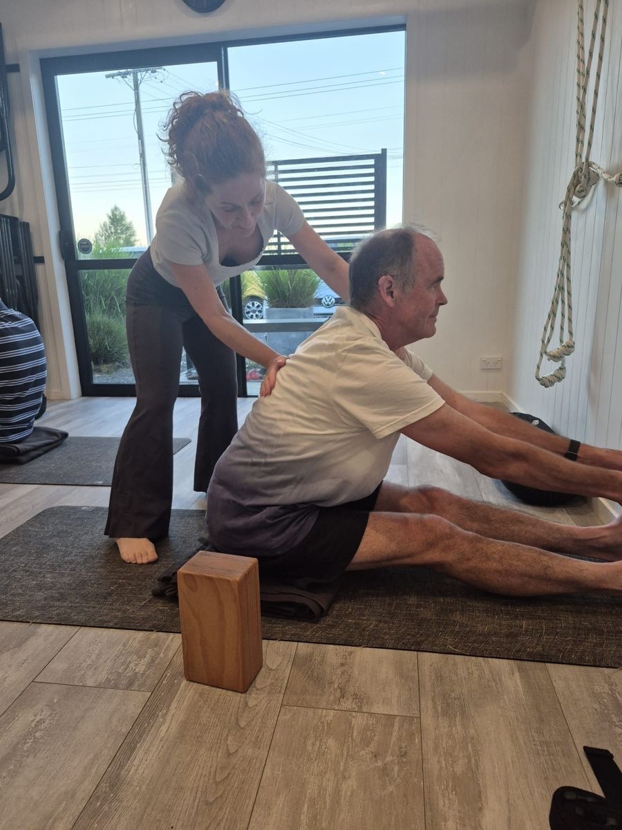 Kathryn giving a student a hands-on adjustment in a seated pose, sitting on a folded blanket with a wooden block beside the mat